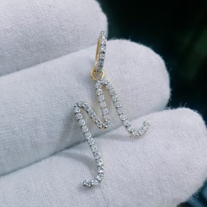 Op de afbeelding: Een goudkleurige oorbel in de vorm van de letter "M", bezet met kleine, sprankelende witte edelstenen. De oorbel wordt getoond tegen een witte achtergrond, wat het gedetailleerde ontwerp en het contrast tussen goud en edelstenen benadrukt.