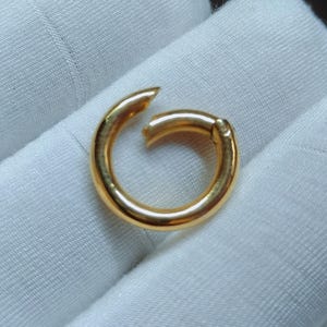 以下が含まれることがあります： 光沢のあるゴールドトーンの円形スプリングリングクラスプ。滑らかで丸みを帯びた金属表面と、小さなヒンジ式の開閉機構が特徴です。このジュエリーパーツは、閉じると継ぎ目のない連続したループのような外観になるように設計されています。