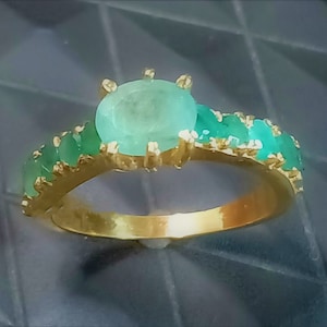 May include: A gold ring featuring a large oval emerald gemstone, surrounded by smaller emerald gemstones.