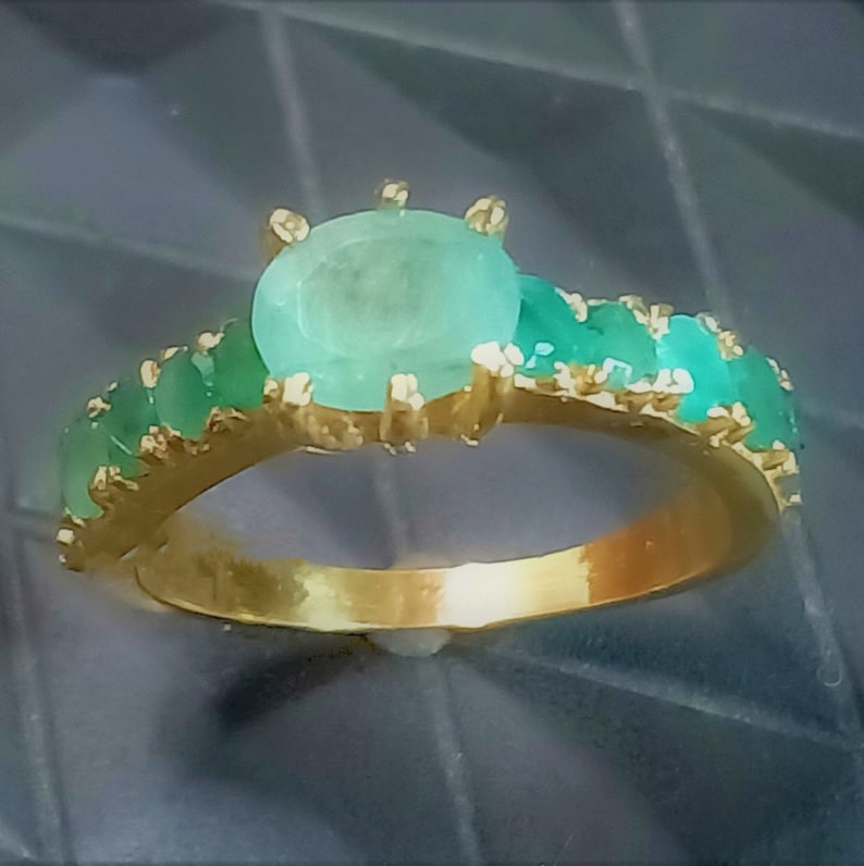 May include: A gold ring featuring a large oval emerald gemstone, surrounded by smaller emerald gemstones.