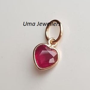 Op de afbeelding: Een hartvormige hanger met een gefacetteerde, dieprode edelsteen gezet in een goudkleurig metaal. De hanger heeft een kleine lus aan de bovenkant om aan een ketting te bevestigen. De tekst "Uma Jewellers" is zichtbaar.