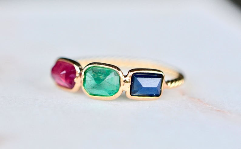 May include: A gold ring with three gemstones: a red ruby, a green emerald, and a blue sapphire.