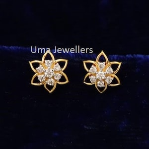 Könnte beinhalten: Ein Paar goldene Ohrstecker in Form von sechsblättrigen Blumen, jeweils mit einer Ansammlung kleiner, klarer Steine in der Mitte. Der Text Uma Jewellers ist über den Ohrringen zu sehen, die vor einem dunkelblauen Samthintergrund liegen.
