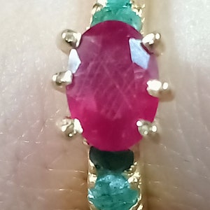 May include: A gold ring featuring a large oval ruby gemstone set within a band of emerald green stones.