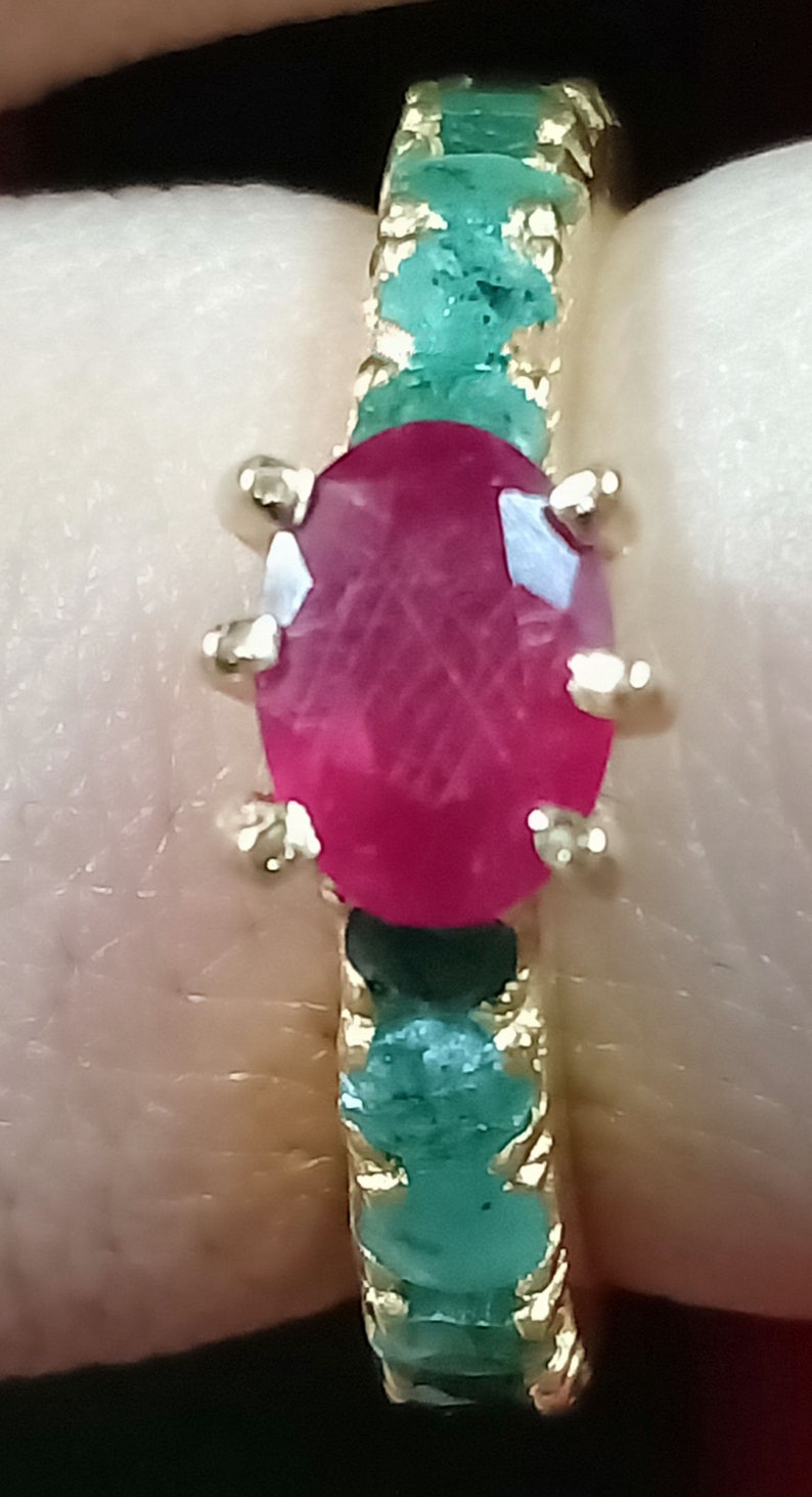May include: A gold ring featuring a large oval ruby gemstone set within a band of emerald green stones.