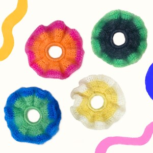 May include: Four colorful, fluffy scrunchies in shades of orange, pink, green, blue, and yellow. Each scrunchie has a ruffled edge and a circular center. The scrunchies are set against a white background with abstract colorful shapes.