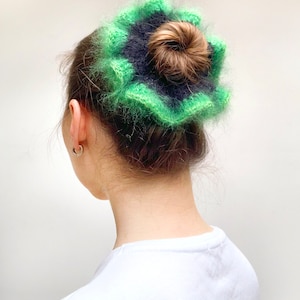 May include: A green and navy blue, fluffy, crocheted hair accessory. The accessory is worn on top of a bun. The hair accessory is a flower shape with a textured, fuzzy appearance. The hair accessory is a unique and stylish hair accessory.