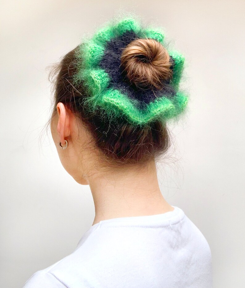 May include: A green and navy blue, fluffy, crocheted hair accessory. The accessory is worn on top of a bun. The hair accessory is a flower shape with a textured, fuzzy appearance. The hair accessory is a unique and stylish hair accessory.