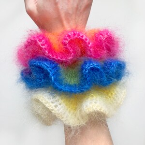 May include: A stack of fluffy, colorful scrunchies in shades of pink, orange, blue, yellow, and green. The scrunchies are made of a soft, fuzzy material and are stacked on top of each other. The scrunchies are a fun and fashionable accessory.