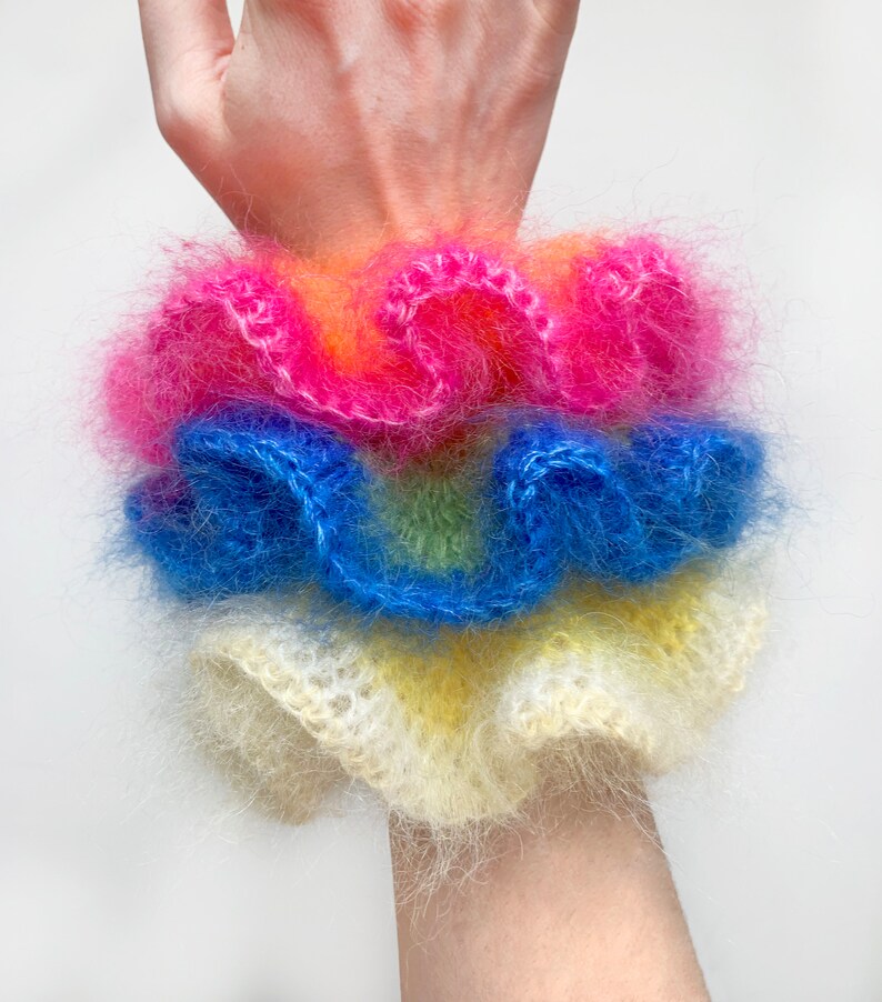 May include: A stack of fluffy, colorful scrunchies in shades of pink, orange, blue, yellow, and green. The scrunchies are made of a soft, fuzzy material and are stacked on top of each other. The scrunchies are a fun and fashionable accessory.