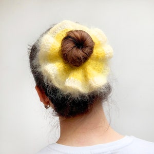 May include: A yellow and white crocheted hair bun cover with a brown hair bun in the center. The hair accessory is made of soft, fluffy yarn and is worn on the back of the head.