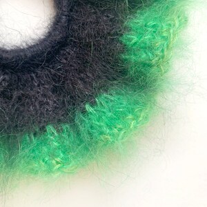 May include: A close-up of a handmade scrunchie with a fluffy texture. The scrunchie features alternating bands of black and bright green yarn. The scrunchie is set against a white background.