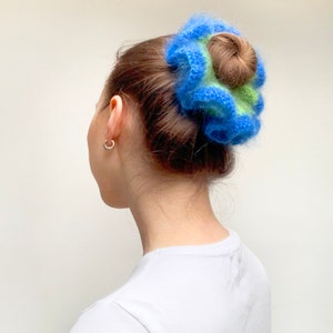 May include: A blue, green, and brown scrunchie. The scrunchie is made of a fluffy material and is wrapped around a bun hairstyle. The person is wearing a white t-shirt and silver hoop earrings.