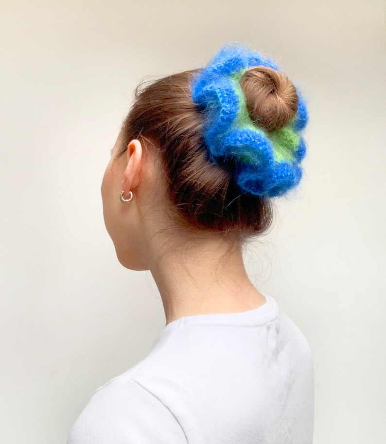 May include: A blue, green, and brown scrunchie. The scrunchie is made of a fluffy material and is wrapped around a bun hairstyle. The person is wearing a white t-shirt and silver hoop earrings.