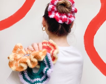 May include: A set of four colorful crocheted hair scrunchies. The scrunchies are in shades of pink, red, orange, yellow, green, and purple. They are all made with a soft, fluffy yarn.