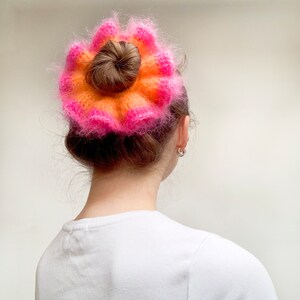May include: A woman's hair is styled in a bun, adorned with a vibrant, fluffy hair accessory. The accessory features a layered design with shades of orange and pink, creating a striking contrast against the dark hair.