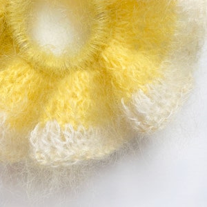 May include: Close-up of a handmade yellow and white scrunchie. The scrunchie is made of a soft, fuzzy material and has a flower-like shape. The color is a bright yellow with white accents. The scrunchie is on a white background.