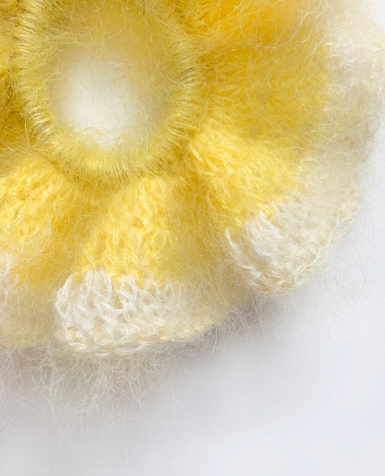 May include: Close-up of a handmade yellow and white scrunchie. The scrunchie is made of a soft, fuzzy material and has a flower-like shape. The color is a bright yellow with white accents. The scrunchie is on a white background.