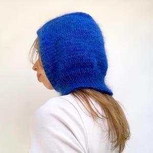Puede incluir: Una balaclava de punto azul vibrante. El gorro está hecho de un hilo suave y texturizado. La balaclava se muestra de lado, con el pelo del usuario y una camiseta blanca visibles.