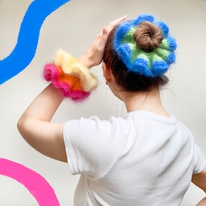 May include: A person wearing a white t-shirt with a colorful, fluffy scrunchie on their wrist and a matching bun wrap. The scrunchie features layers of pink, orange, and yellow. The bun wrap is blue, green, and brown.