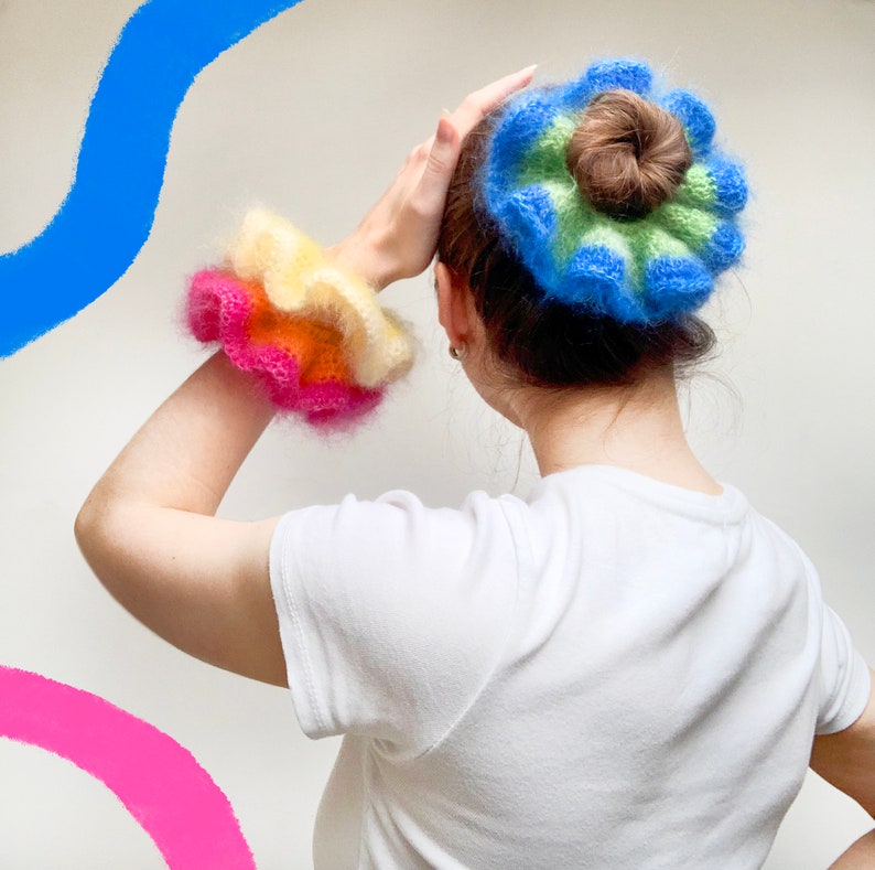 May include: A person wearing a white t-shirt with a colorful, fluffy scrunchie on their wrist and a matching bun wrap. The scrunchie features layers of pink, orange, and yellow. The bun wrap is blue, green, and brown.