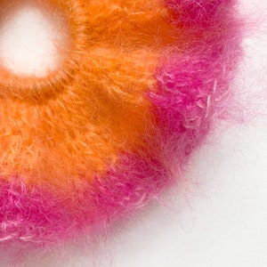 May include: Close-up of a fluffy scrunchie in shades of orange and pink. The scrunchie is made of a soft, textured material, perfect for adding a pop of color to any hairstyle. The vibrant colors create a playful and eye-catching accessory.