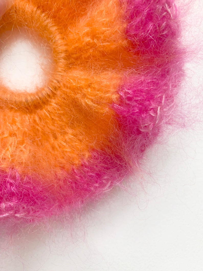 May include: Close-up of a fluffy scrunchie in shades of orange and pink. The scrunchie is made of a soft, textured material, perfect for adding a pop of color to any hairstyle. The vibrant colors create a playful and eye-catching accessory.
