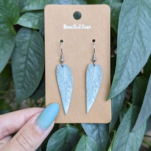 May include: A pair of silver leaf earrings with a light blue, veined design. The earrings are on a brown card with the text "Born Sick Sage".