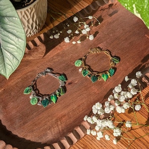 Puede incluir: Dos pulseras con dijes de hojas. Una es plateada, la otra dorada. Ambas presentan dijes verdes en forma de hoja con diferentes tonos de verde y detalles blancos. Las pulseras se exhiben en una bandeja de madera con flores blancas.
