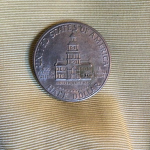 Könnte beinhalten: Eine silberne Halbdollar-Münze mit der Aufschrift "United States of America" und "200 Years of Freedom" am Rand. Die Mitte der Münze zeigt eine Darstellung der Independence Hall mit der Aufschrift "Independence Hall" darunter.