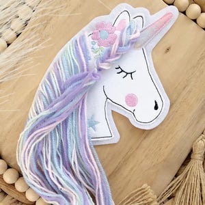 Könnte beinhalten: Ein skurriles Einhorn-Applikation mit weißem Gesicht, rosa Wangen und geschlossenen Augen. Das Einhorn hat ein rosa Horn und einen Blumenkranz. Seine fließende Mähne besteht aus pastellfarbenem Garn. Die Applikation befindet sich auf einer gewebten Holzbasis.