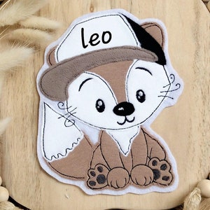 Könnte beinhalten: Ein niedlicher, gestickter Flicken mit einem Fuchs, der eine Baseballkappe mit dem Namen "Leo" trägt. Der Fuchs ist hauptsächlich braun und weiß, mit schwarzen Akzenten. Der Flicken befindet sich auf einer Holzoberfläche, umgeben von einem geflochtenen Korb und Holzperlen.