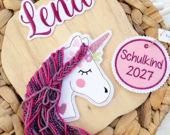 Könnte beinhalten: Ein weißer Einhorn-Aufnäher mit glitzerndem Horn, rosa Wangen und einer langen Mähne aus violetter und rosa Wolle. Darüber befindet sich ein Aufnäher mit dem Namen Lena in magentafarbener Schrift. Rechts ein runder rosa Aufnäher mit Schulkind 2027.