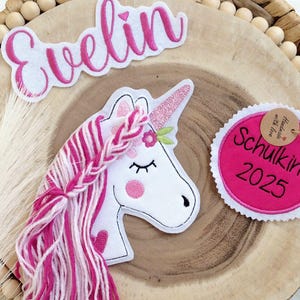 Könnte beinhalten: Ein skurriles Einhorn-Patch mit rosa und weißer Mähne, einem glitzernden Horn und einem floralen Akzent. Der Name "Evelin" ist über dem Einhorn gestickt. Ein rosa runder Patch mit dem Text "Schulkind 2025" ist ebenfalls vorhanden. Die Patches befinden sich auf einer Holzscheibe.