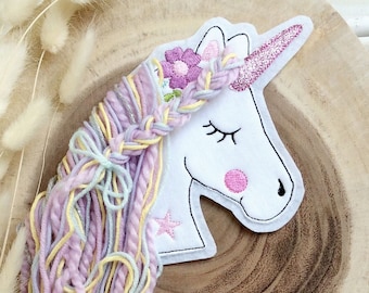 May include: A white unicorn head with a pink glitter horn, closed eyes, and a pink cheek. The unicorn has a braided mane with a flower crown and long, flowing yarn hair in pastel colors of pink, yellow, and blue. The unicorn is on a wooden surface.