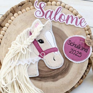 Könnte beinhalten: Dekorative Holzplakette mit einem Pferdekopf-Design und langer, fließender Mähne. Das Pferd hat ein rosa Halfter und den Namen "Salome" darüber. Ein Etikett lautet "Schulkind 2025". Die Plakette befindet sich auf einer gewebten Basis mit Holzperlen.
