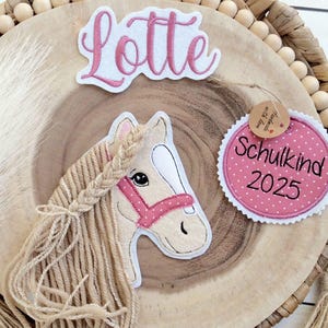 Könnte beinhalten: Dekorative Holzplakette mit Pferdeapplikation, dem Namen "Lotte" und einem Anhänger mit der Aufschrift "Schulkind 2025". Das Pferd hat eine geflochtene Mähne und einen rosa Zaum. Die Plakette ist von gewebtem Material und Holzperlen umgeben.