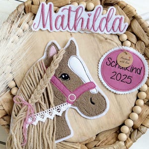 Könnte beinhalten: Wanddekoration mit einem aufgestickten Pferdekopf, geflochtener Mähne und rosa Halfter. Der Name "Mathilda" steht über dem Pferd, und ein rosa Etikett zeigt "Schulkind 2025". Der Hintergrund ist ein geflochtener Korb.