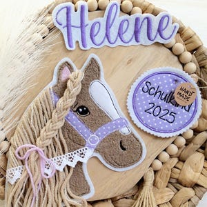 Könnte beinhalten: Ein dekorativer Pferdekopf mit geflochtener Mähne, lila Zaumzeug und dem Namen "Helene" in lila Schrift. Ein kreisförmiges Etikett lautet "Schulkind 2025" und "Hand Made". Das Pferd befindet sich auf einer gewebten Holzbasis mit Holzperlen.