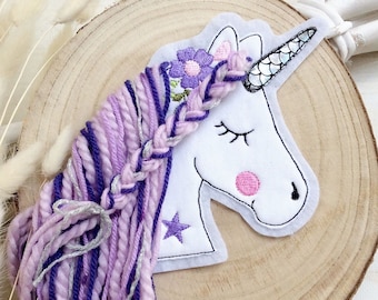 Könnte beinhalten: Ein weißer Einhornkopf mit geschlossenen Augen, rosa Wangen und einem schwarzen Horn. Das Einhorn hat eine geflochtene Mähne in Lila und Silber sowie einen lila Blumenkranz. Das Einhorn liegt auf einer Holzscheibe.