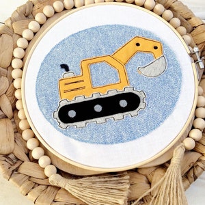 Könnte beinhalten: Gesticktes Hoop-Art-Stück mit einem gelb-schwarzen Cartoon-Bagger auf hellblauem Hintergrund. Die Stickerei befindet sich in einem Holzreifen und wird auf einem gewebten Tablett mit Holzperlen und einem Quastenanhänger präsentiert.