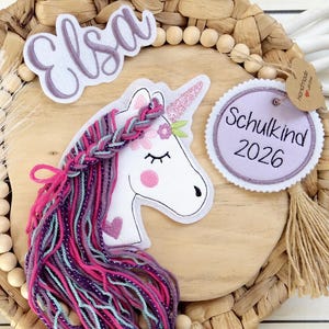 Könnte beinhalten: Dekorative Holzplakette mit Einhorn-Design, dem Namen "Elsa" und dem Text "Schulkind 2026". Das Einhorn hat eine rosa und lila Mähne, ein glitzerndes Horn und befindet sich auf einem gewebten Hintergrund mit Holzperlen.