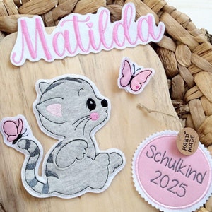Könnte beinhalten: Holzschild mit grauem Katzen-Applikation, rosa Schmetterlingen und dem Namen "Matilda" in Rosa. Ein rosa-weißer Aufnäher zeigt "Schulkind 2025". Das Schild befindet sich vor einem geflochtenen Hintergrund.