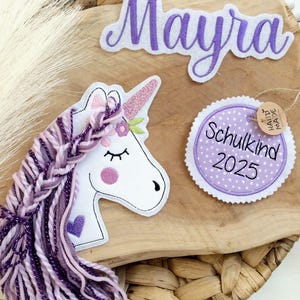 Könnte beinhalten: Ein Einhorn-Set mit einem weißen Einhornkopf mit geflochtener lila Mähne, einem Namensschild mit der Aufschrift "Mayra" in lila Schrift und einem runden Etikett mit der Aufschrift "Schulkind 2025". Die Artikel sind auf einer Holzoberfläche angeordnet.