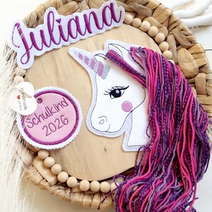 Könnte beinhalten: Ein Wandbehang mit einem weißen Einhornkopf, einem silbernen Horn und einer langen Mähne aus rosa und violettem Garn. Das Stück enthält ein Namensschild mit der Aufschrift Juliana, einen runden rosa Aufnäher mit dem Text Schulkind 2026 und Holzperlen.