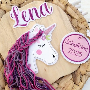 Könnte beinhalten: Holzschild mit Einhorn-Design, dem Namen "Lena" und einem Anhänger mit der Aufschrift "Schulkind 2025". Das Einhorn hat ein rosa Glitzerhorn, rosa Wangen und eine geflochtene Mähne aus rosa und lila Garn.