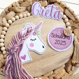 Könnte beinhalten: Eine verspielte Anordnung mit einer Holzbasis, einem Einhorn-Applikation, dem Namen "Greta" und einem "Schulkind 2025"-Abzeichen. Das Einhorn hat ein rosa Horn und eine geflochtene Mähne aus rosa und lila Garn. Die Anordnung befindet sich auf einer geflochtenen Weidenbasis.