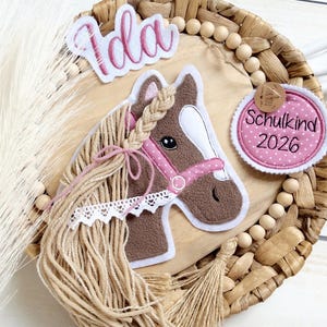 Könnte beinhalten: Ein dekoratives Pferdekopf-Design mit dem Namen "Ida" in rosa Schrift. Das Pferd hat einen braunen Kopf, eine rosa Trense und eine geflochtene Mähne. Ein kreisförmiger Aufnäher zeigt "Schulkind 2026" in Rosa. Das Design befindet sich auf einem gewebten Hintergrund.