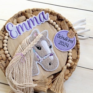 Könnte beinhalten: Wanddekoration mit einem Pferdekopfabzeichen und einer langen, geflochtenen Mähne. Der Name "Emma" ist über dem Pferd gestickt. Ein kreisförmiger Aufnäher lautet "Schulkind 2024". Das Ganze befindet sich auf einer gewebten Basis mit Holzperlen.