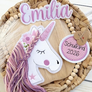 Könnte beinhalten: Dekoration mit Einhorn-Motiv, mit einem weißen Einhornkopf, einem rosa Glitzerhorn und einer geflochtenen Mähne aus rosa und violettem Garn. Der Name "Emilia" steht über dem Einhorn, und ein Etikett lautet "Schulkind 2026".